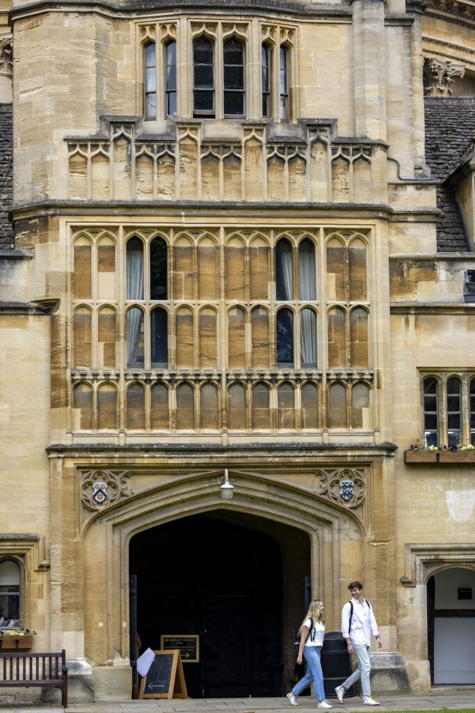 A male and a female college student walk and talk together with a large, ornate stone building in the background.