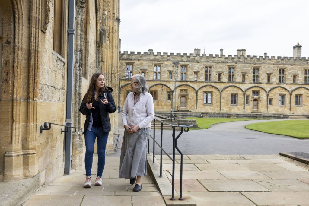 A female college student walks with a silver-haired woman on a stone ramp. It’s a gray, overcast day and sandstone walls that almost look like part of a castle are behind them.