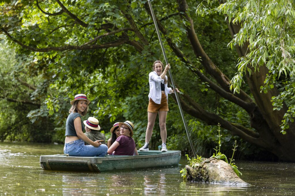 Four young women wearing straw boater hats sit in a long narrow boat, and another one, in mid-laugh, stands holding a pole into the water.