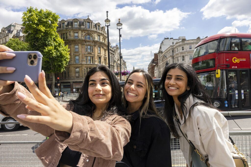 Three students pose for a selfie with a double-decker red bus in the background.