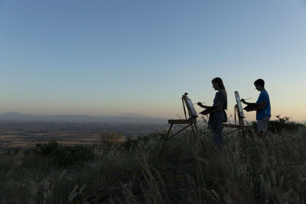 A student paints on a canvas atop a tripod. He is in the middle of a grassy field.