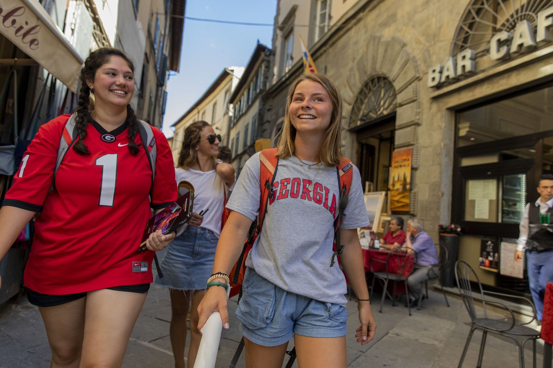 A female student is wearing jean shorts and a grey shirt with the University of Georgia logo. She happily walks through the streets of a European city.