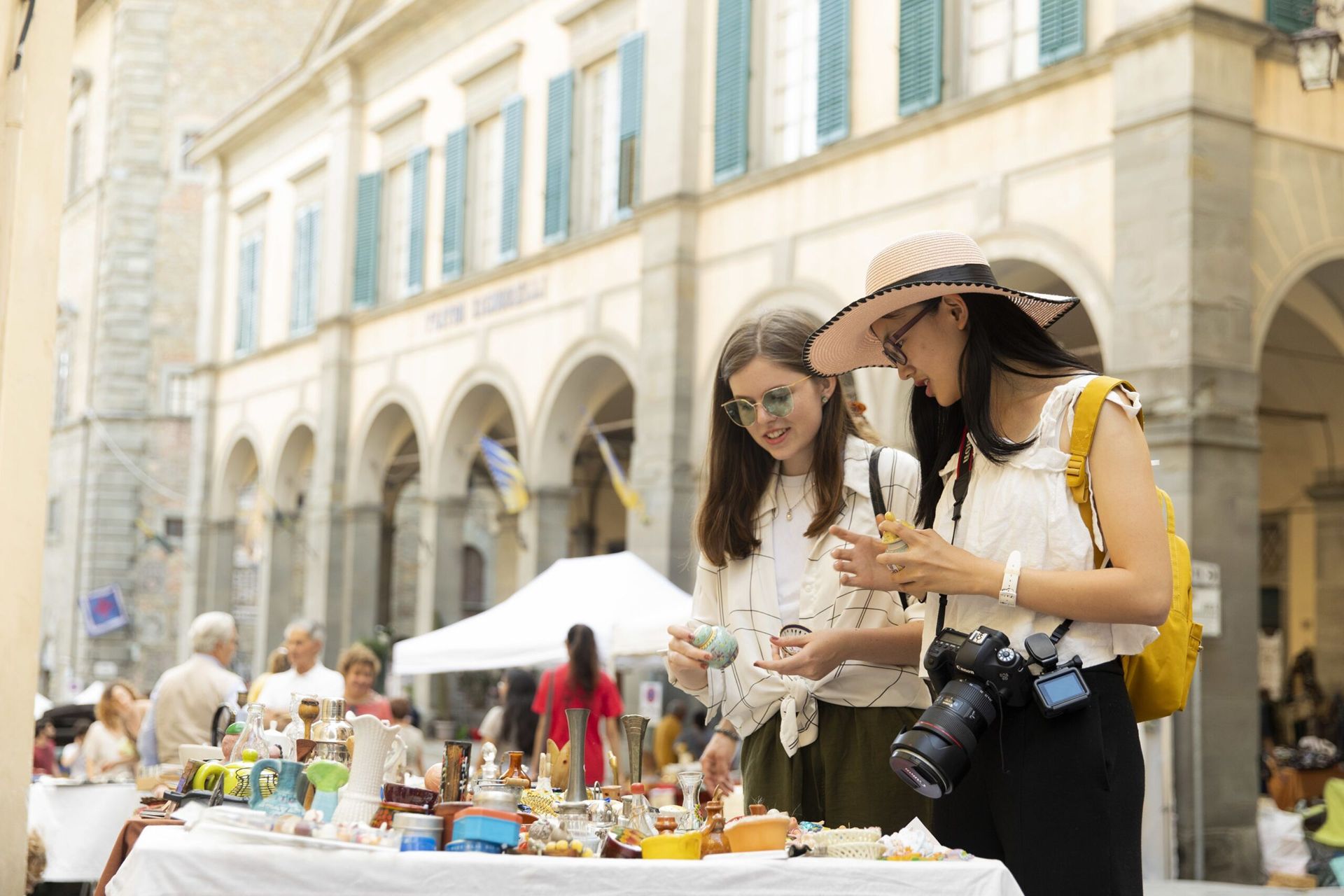 Two women in white tops and dark bottoms look at wares in a street market.