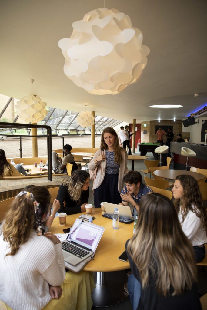 A group of students sits around a round wooden table as they study and talk. They are in a large, modern room with other students.