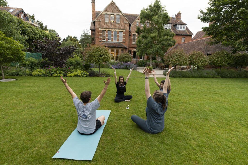 Three students exercise on a bright green lawn. They are sitting in a circle and each has their hands raised to the sky.