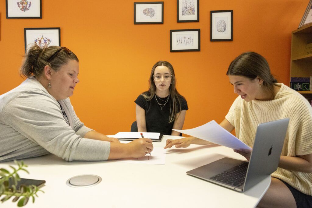 In a small starkly orange office, a female undergraduate student points to a white sheet of paper that a female professor is marking on with a pencil. Another female student looks on.