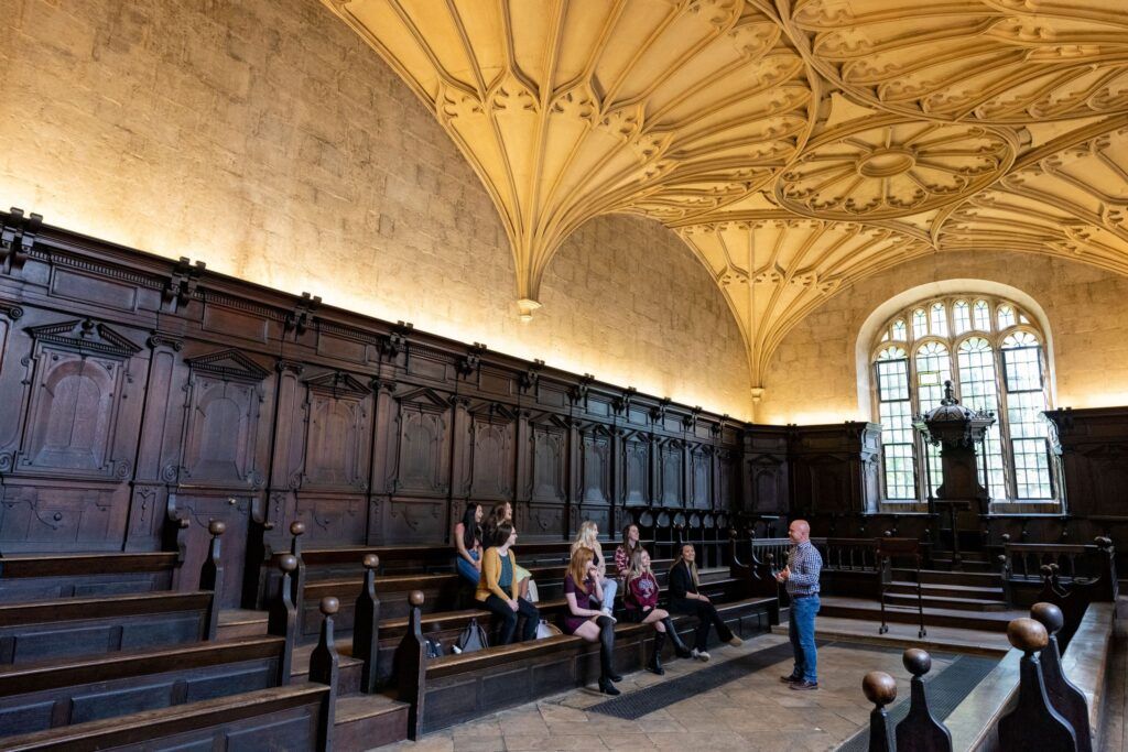 A man stands and speaks to smiling female students sitting on pews in a stately room with dark wooden paneling, a stone floor, and an ornately carved stone ceiling.