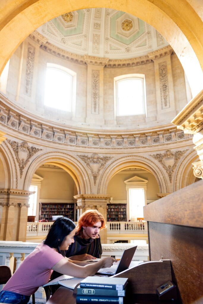 Two students sit in a beautiful, bright library looking at a laptop screen. In the background is a magnificently designed interior dome ceiling of gray, white, and turquoise.