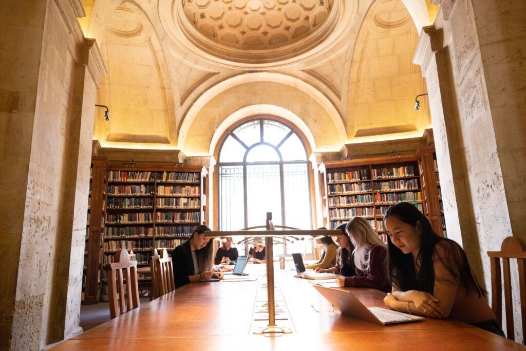 Six students sit at an extended library desk looking at their laptops or study materials with the sun shining through a window in the background.