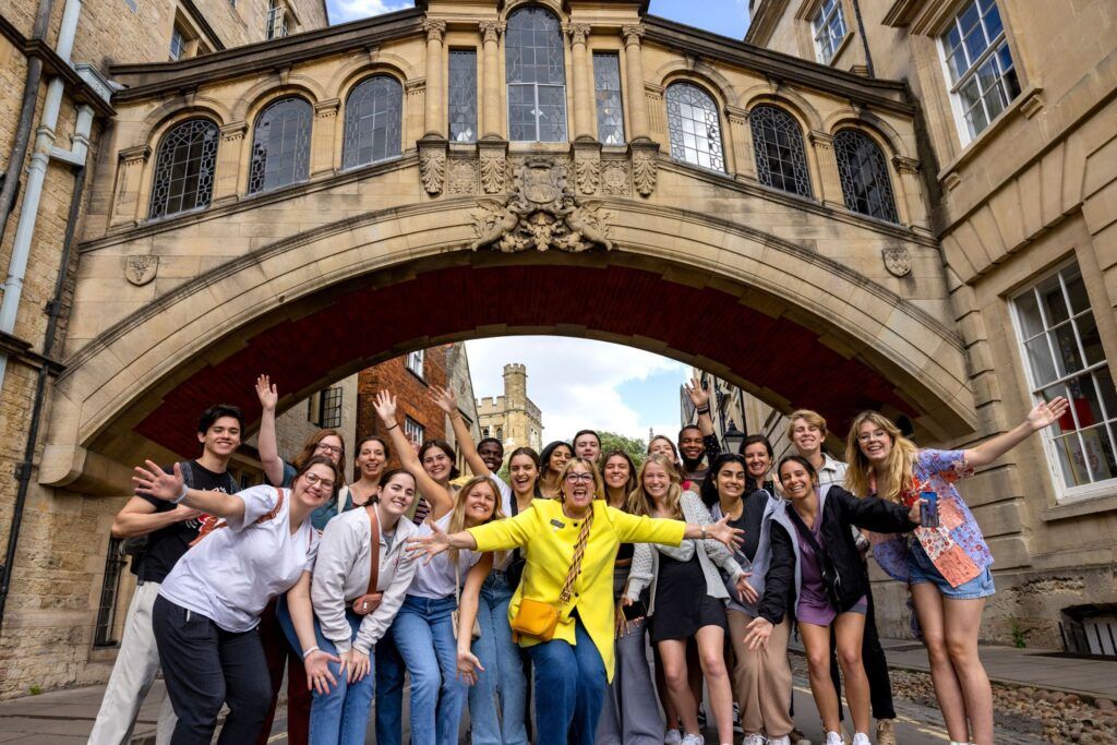 About a dozen students pose for a photo with a woman in a yellow jacket, at the center, spreading her arms. They are under an elevated stone walking bridge that connects two buildings.