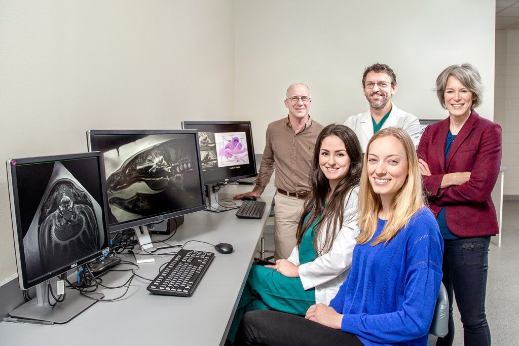 A group of five people smile at the camera from a medical office from the right side of the photo. On the left of the photo are three computer screens all showing MRI images.