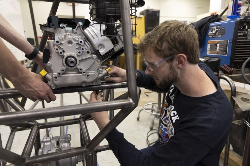 A bearded man wearing safety goggles and a long-sleeved black shirt works on part of a metal frame that is the back end of a Baja vehicle under construction.