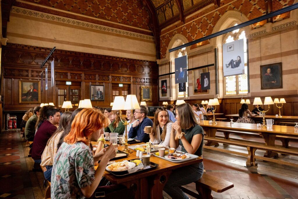 Students sit at a long table sipping coffee and eating food in a room with vaulted ceilings, wood-panel and tiled walls, and large arched windows.