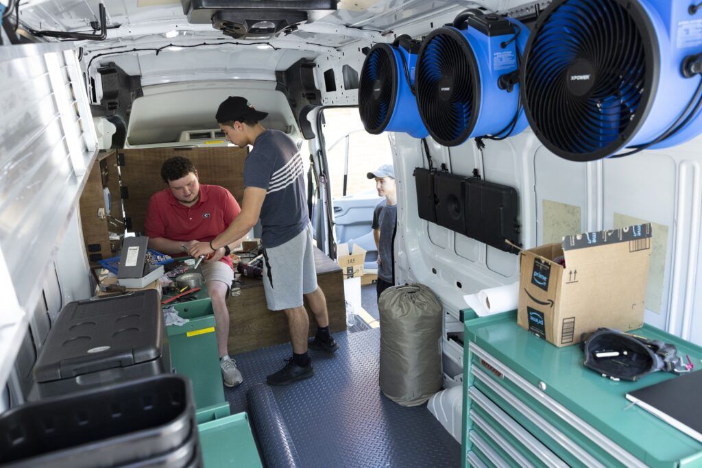 Two university students work on an engineering project from the inside of a white van. The van has three blue vans along the top of one wall and several green-blue storage cabinets.