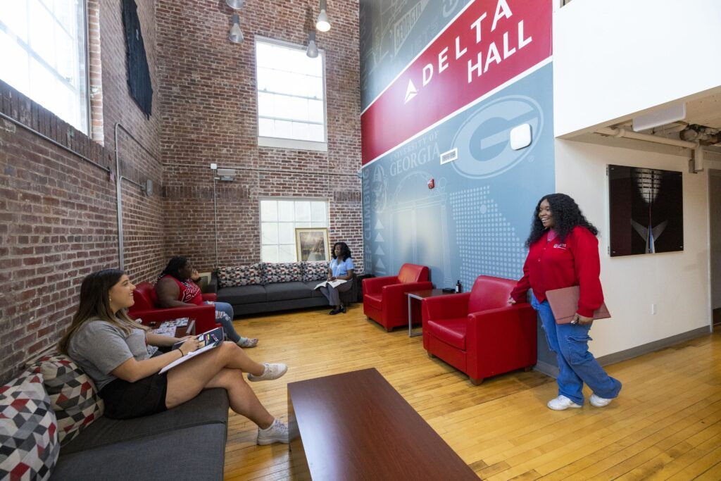 Students chat in a common area on their college. The space has a long dark couch along the brick wall, a couple of comfortable looking red chairs, and a short brown table.