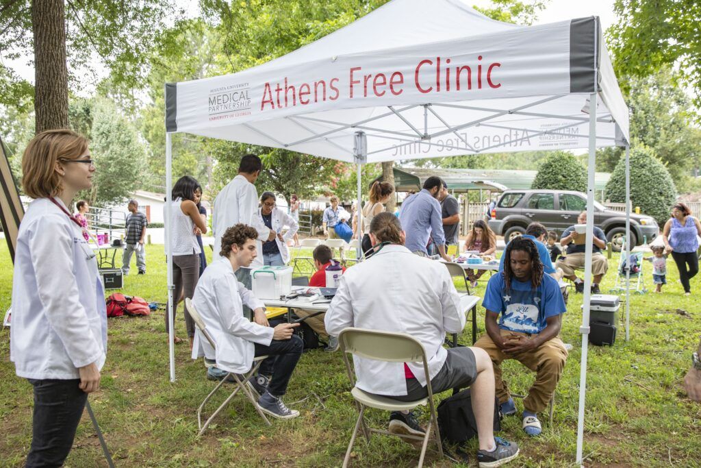 Several medical students and patients interact under a white tent 
that has the text “Athens Free Clinic”. The tent is outside on the grass and 
there are many other people in the background.