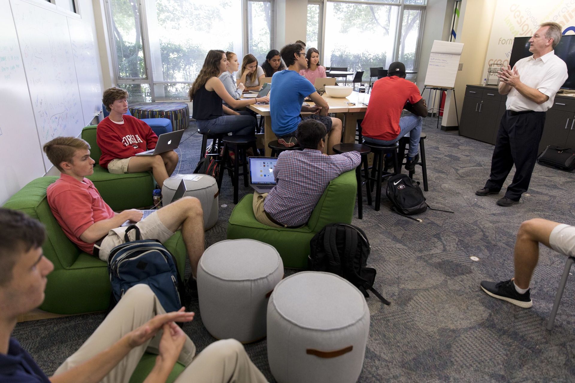 A large group of students sit in a classroom and listen to a lecturer. The lecturer is wearing dark pants and a white shirt. The students are in various positions. Some sit on short green fabric chairs and others sit around a tall brown table.