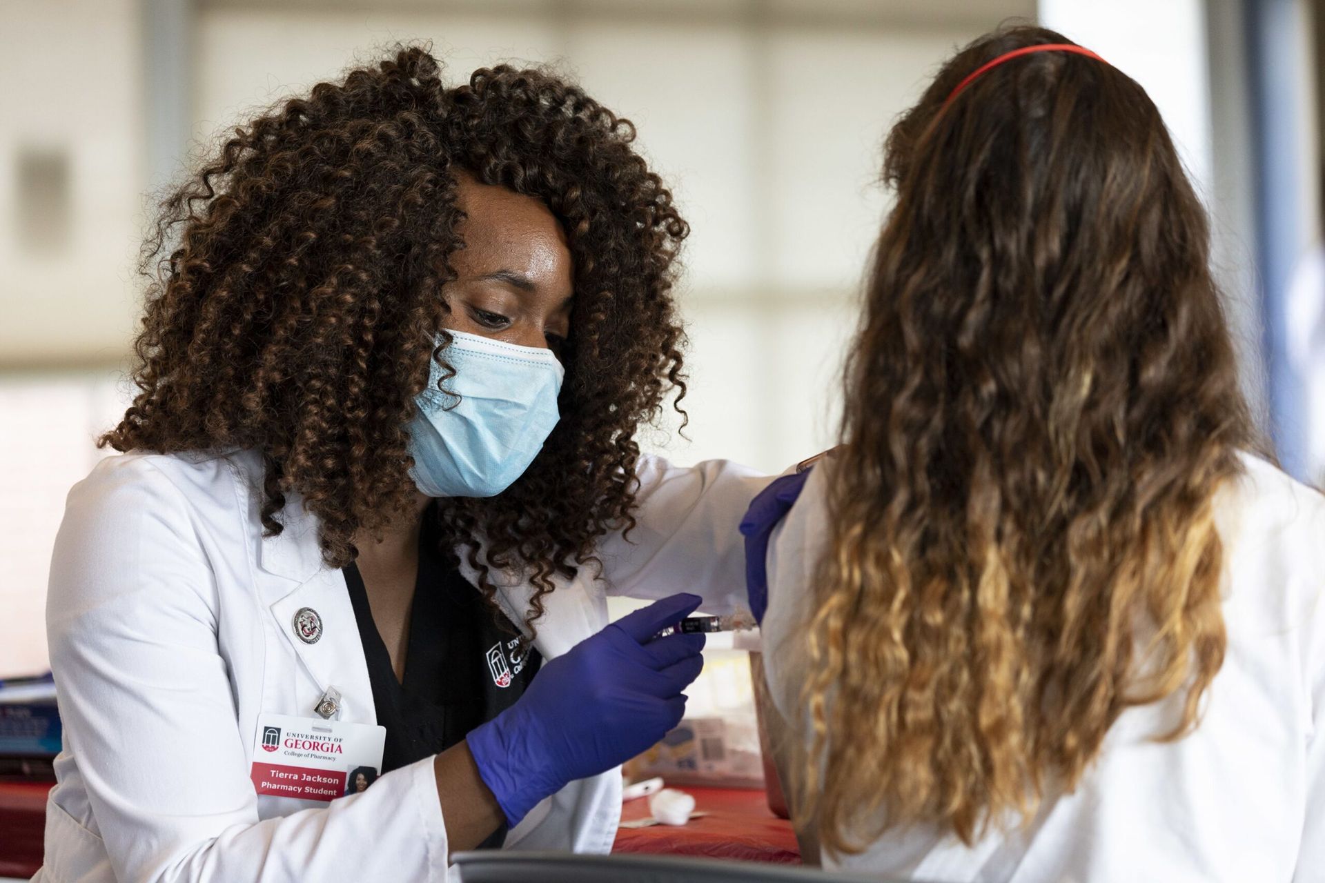 A medical student wears a white lab coat, blue medical face mask, dark blue medical gloves, and University of Georgia badge. She gives a shot to a patient who is facing away from the camera. The patient wears a white shirt and a red headband.