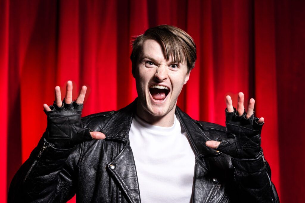 A chest-up photo of an actor dressed as Orin in Little Shop of Horrors. His mouth is wide open and his open palms are held up at shoulder level.