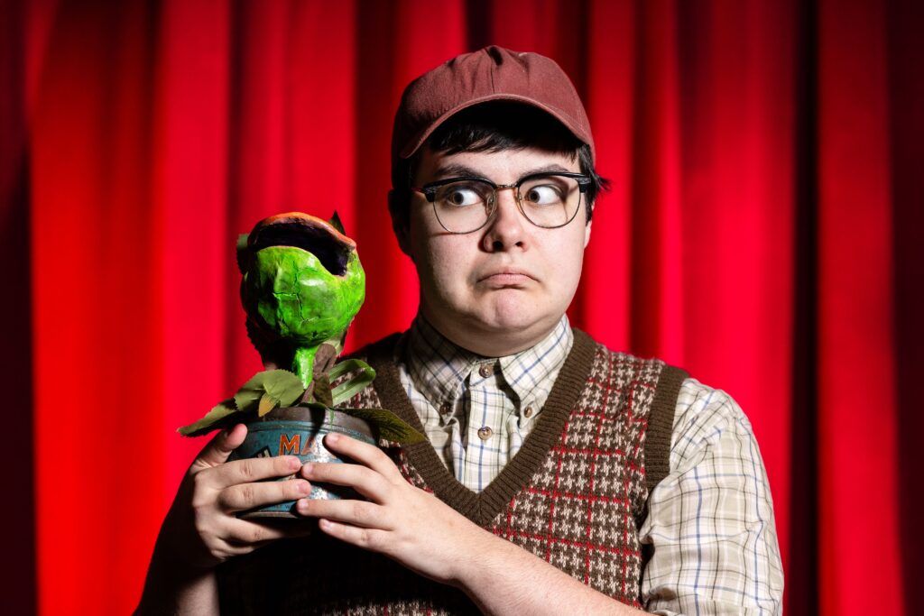 A chest-up photo of an actor dressed as Seymour in Little Shop of Horrors. He is smiling at the camera and holding a fake plant.