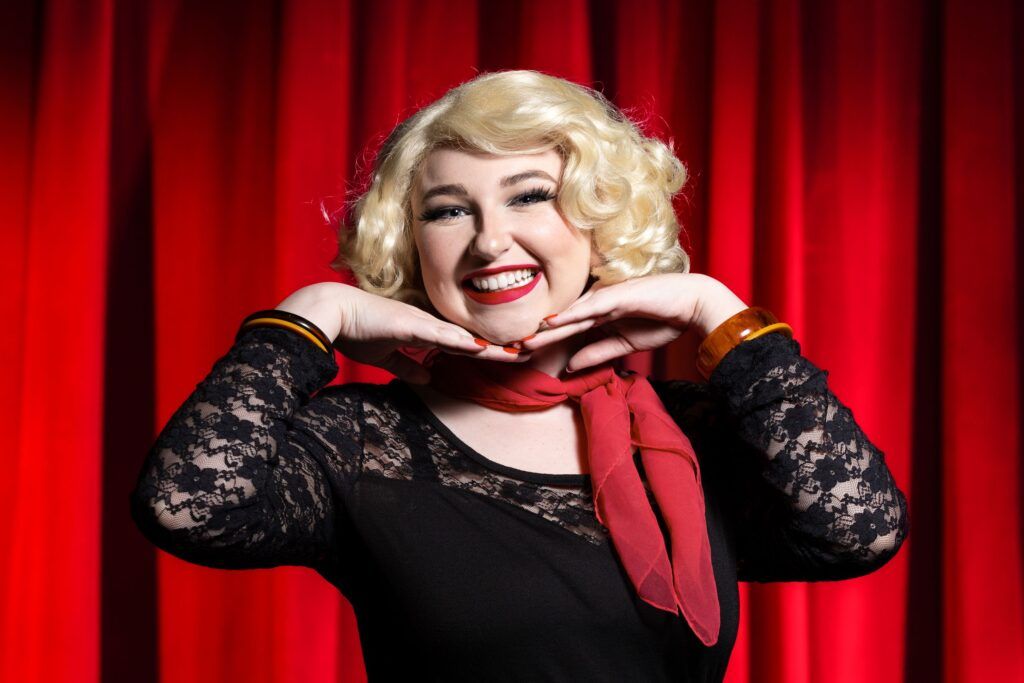 A chest-up photo of an actor dressed as Audrey in Little Shop of Horrors. She is smiling at the camera and posing with her hands under her chin.