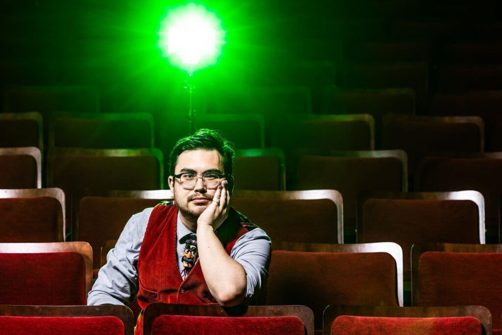 A young man in a blue shirt and red vest sits in a darkened theatre. He stares at the camera with one hand on his face and that elbow on a theatre seat. There is a bright green light shining behind him.