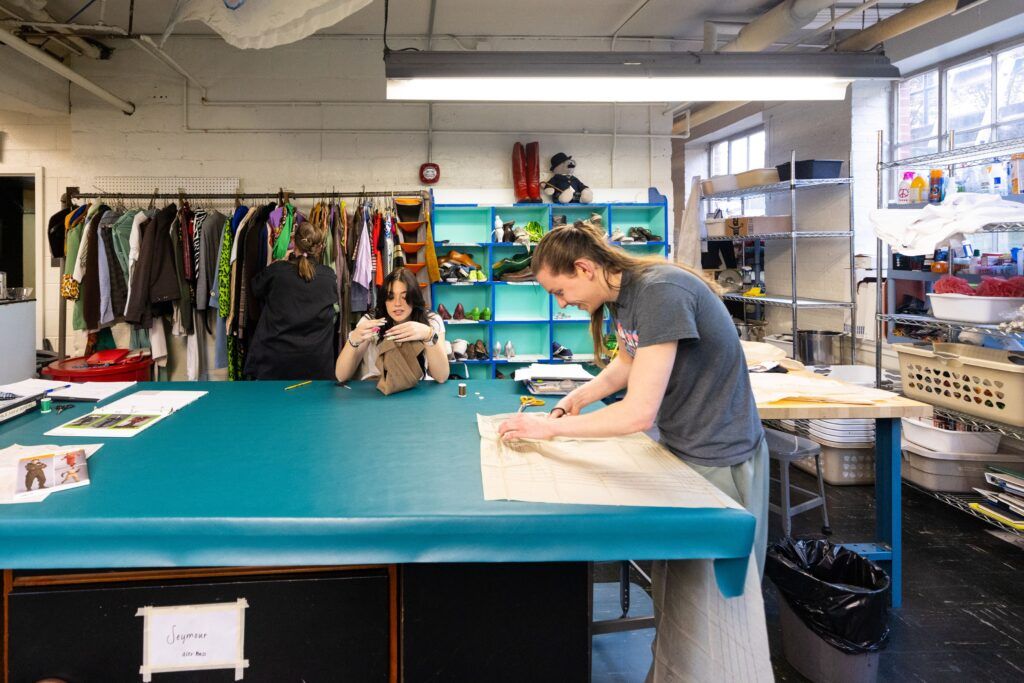 Two students from the UGA theatre department work on clothing design for the Little Shop of Horrors. The stand at a large blue table and are surrounded by racks of clothing and containers of sewing materials.