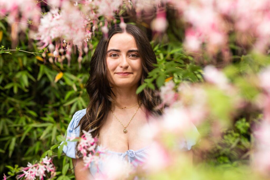 An artistic photo of a young woman, Abby McWethy, as she looks at the camera from behind several branches with bright pink flowers. The photo is slightly blurred.