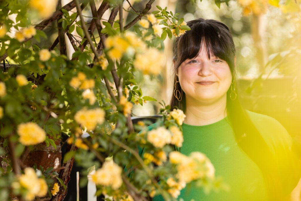 An artistic photo of a young woman, Sloane Elle Garner, as she looks at the camera from behind several branches with bright yellow flowers. The photo is slightly blurred.