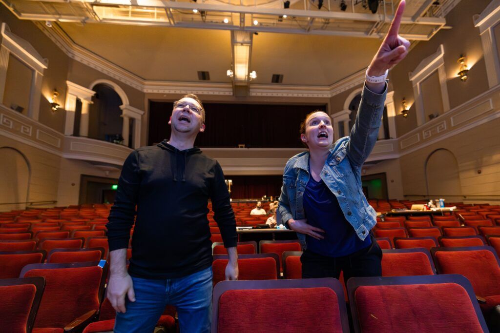 A man and a woman stand behind a row of theatre seats and give direction towards the stage.