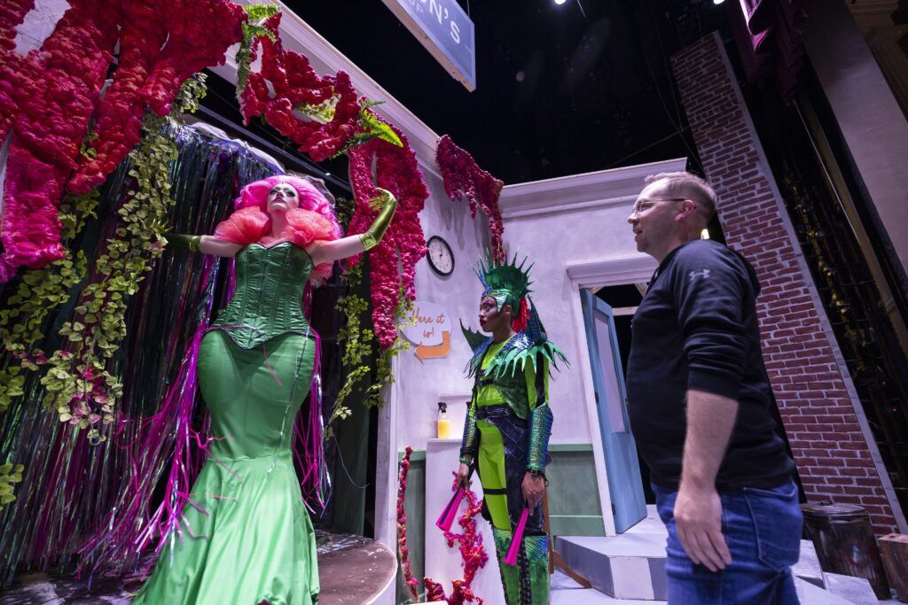 A director gives notes to an actor playing Audrey 2 in a performance of Little Shop of Horrors. The actor is in full costume with a long, tight green dress and bright pink hair. They are surrounded by fake pink and green hanging plants. 