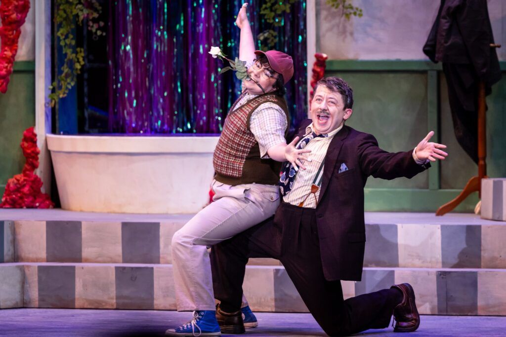 Two male actors perform on stage as Seymour and Mr. Mushnik during a performance of Little Shop of Horrors. One actor is wearing a red cap and patterned vest as he sits on the knee of the other actor who is doing a short lunge on the floor.