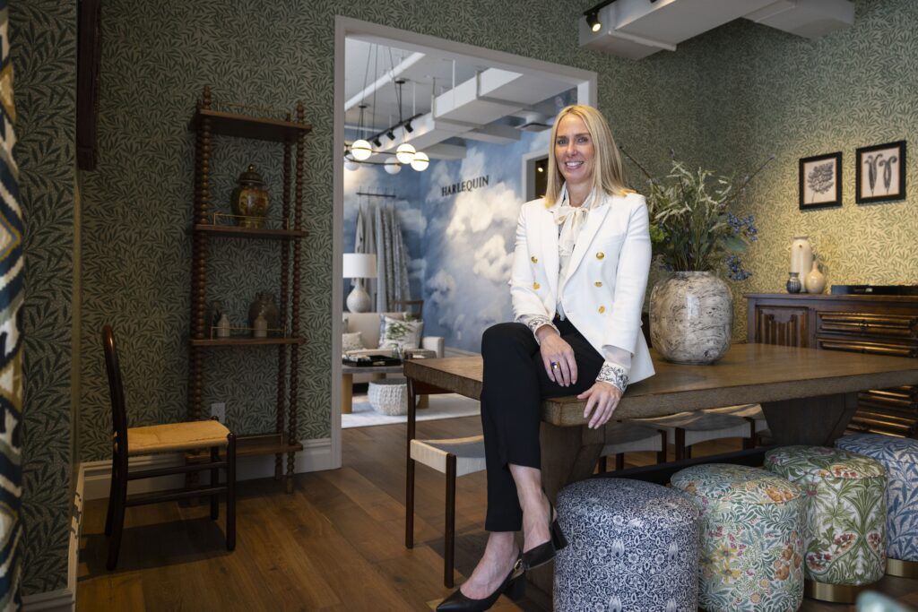 A woman in black pants and a white jacket sits cross-legged on a wooden table. The room is decorated in a variety of blue and green patterned objects and wallpaper.