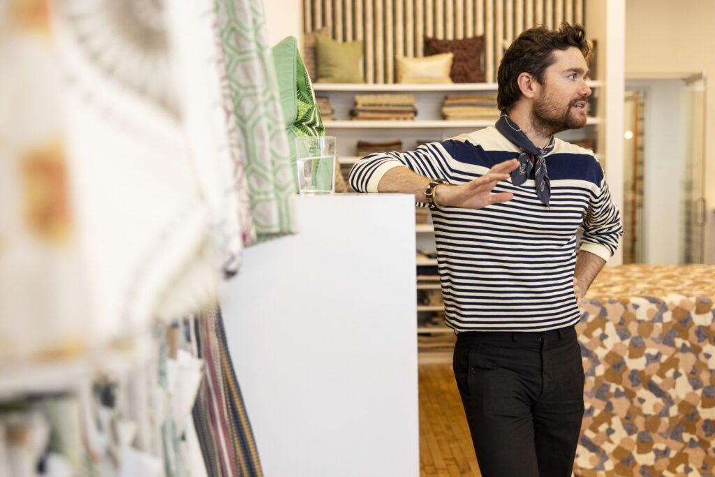 A man in a blue and white striped shirt and blue patterned neckerchief leans against a white shelf while speaking to someone off-camera. There are several different types of fabric hanging on the left side of the photo and a shelf full of folded fabrics and pillows in the background.