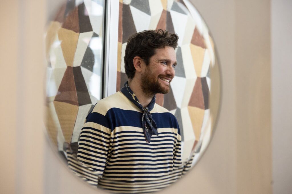 A closeup of a smiling young man reflected in a round mirror. He is wearing a blue and white striped shirt with a blue neckerchief.