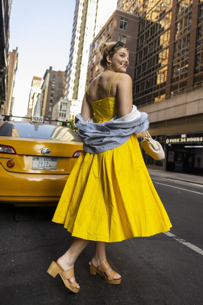 A young woman in a yellow dress and heeled sandals walks past a cab while turning her head to smile behind her.