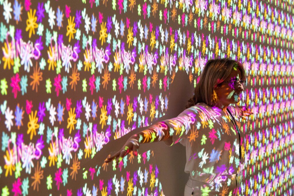 A young woman stands with her arms outstretched against a wall. Different colored ribbons and bows are projected across her and the wall.