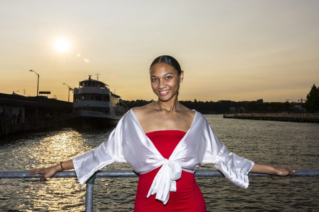 A closeup of a young woman facing forward against the railing of a boat. She is wearing a red strapless dress with a white top tied around her. There is a dock and boat in the background.