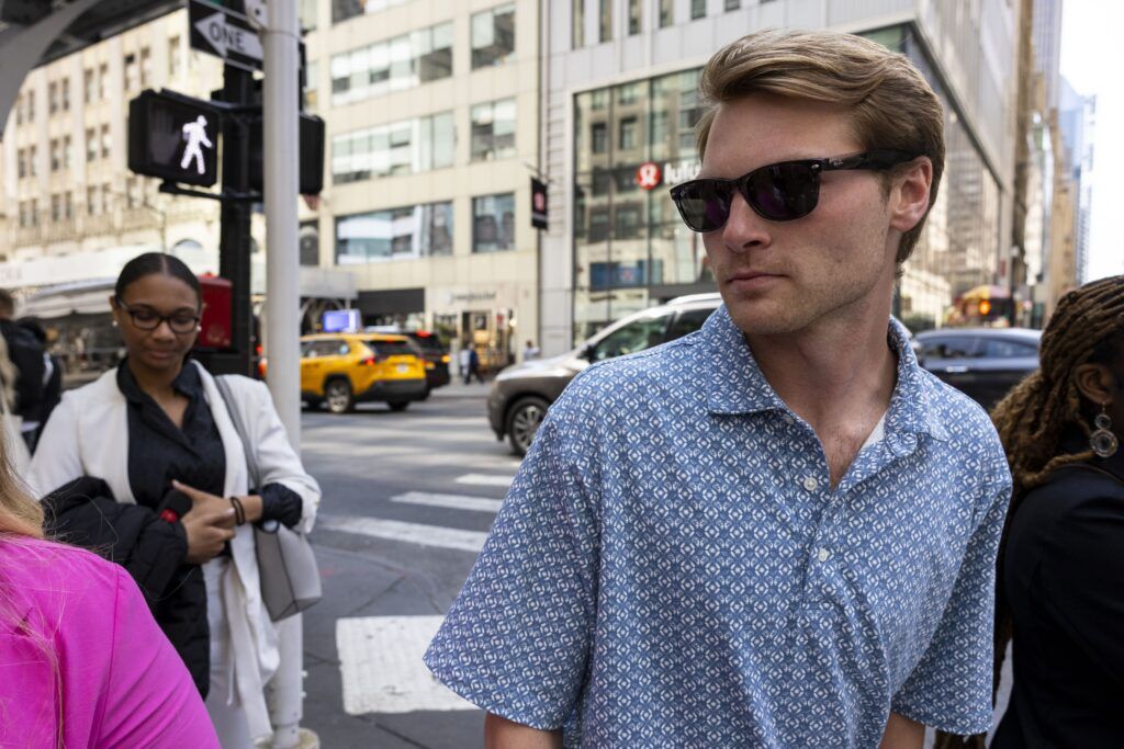 A young man in a blue and white patterned shirt and sunglasses crosses the street in a busy city. There are several other people around him and light-colored buildings in the background.