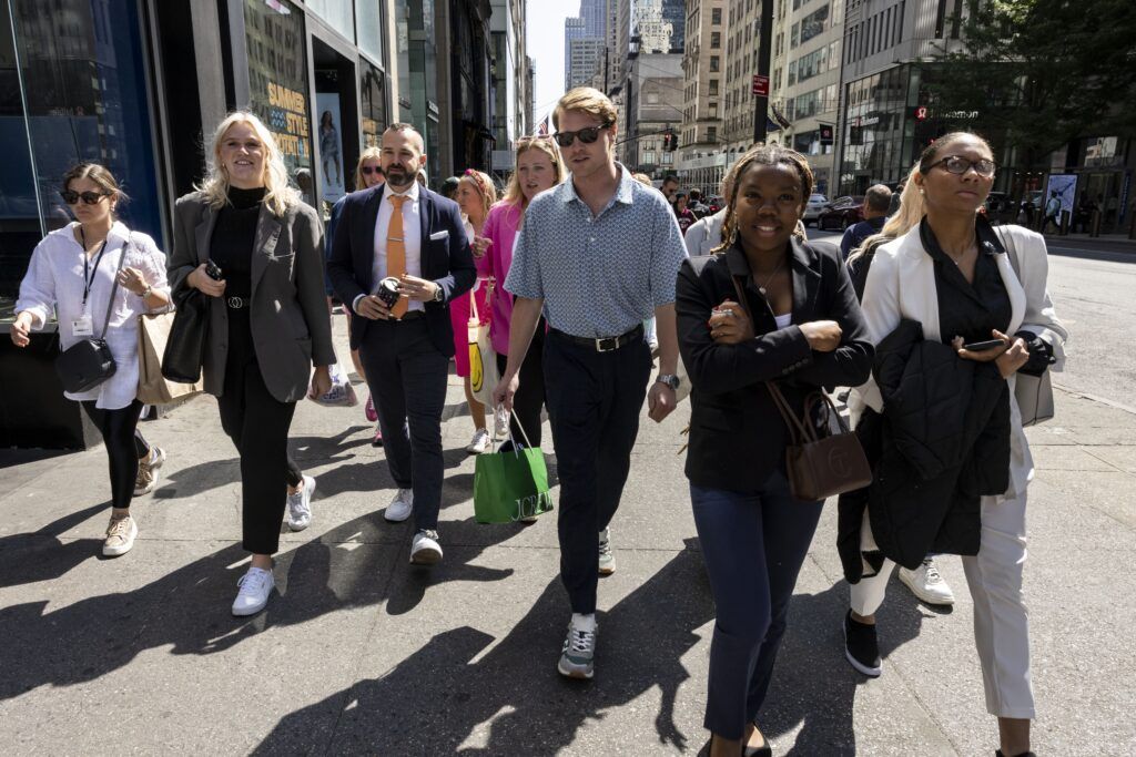 A crowd of people dressed in business casual clothing walk forward on the street. In the background are several modern buildings with glass windows.