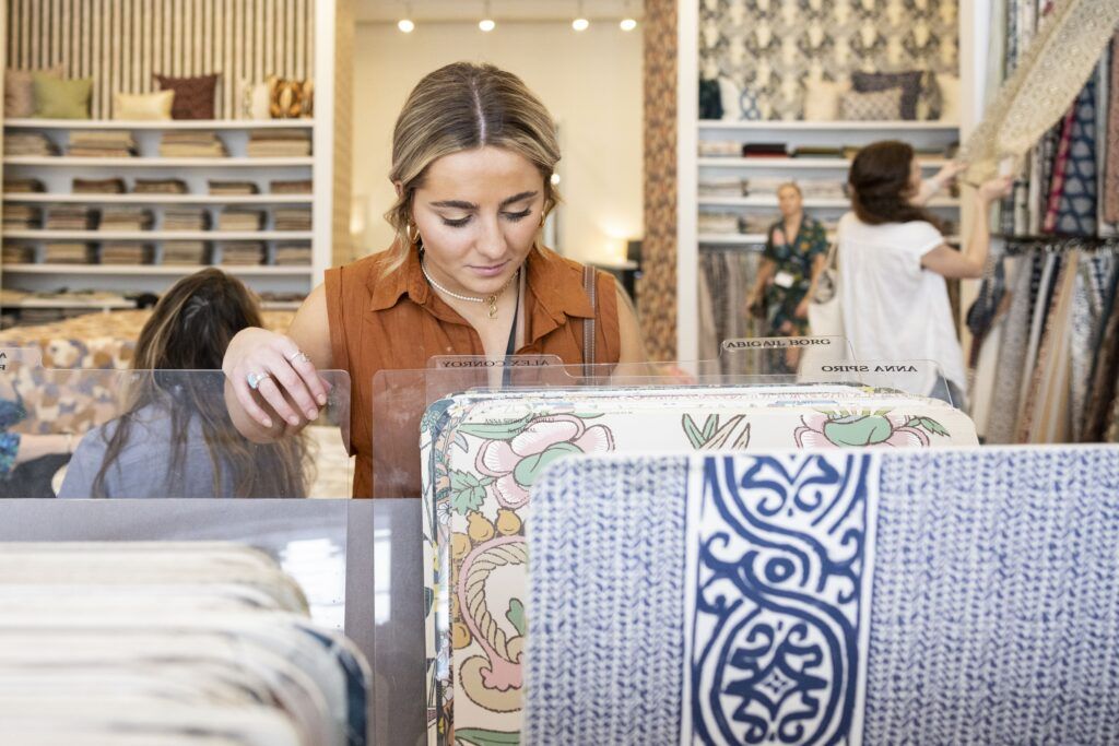 A young woman in an orange shirt looks through fabric options. In the background, several other young women do the same.