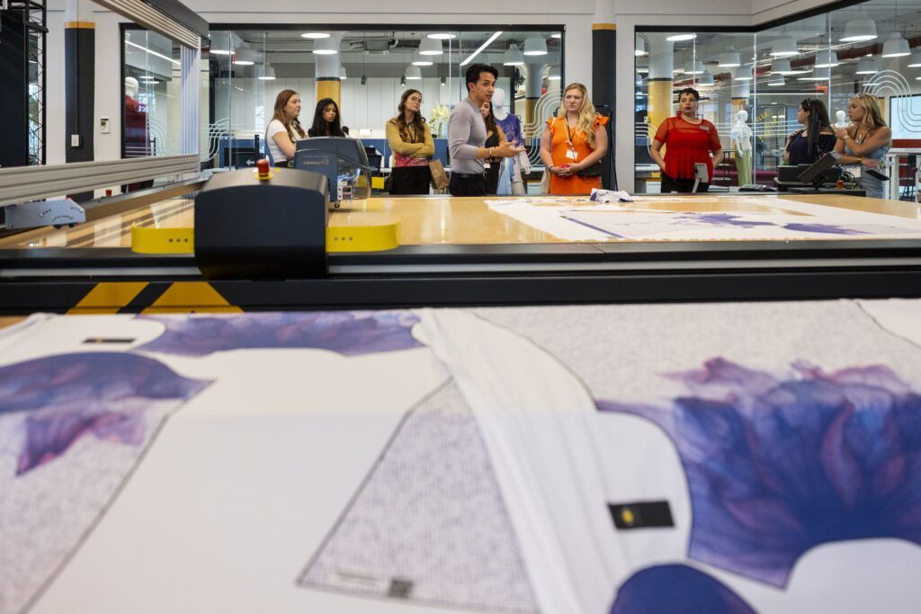 Several university students learn about fashion merchandising and business in a clothing factory. There is a large wooden table in the center of the photo and the students and tour guide stand behind it.