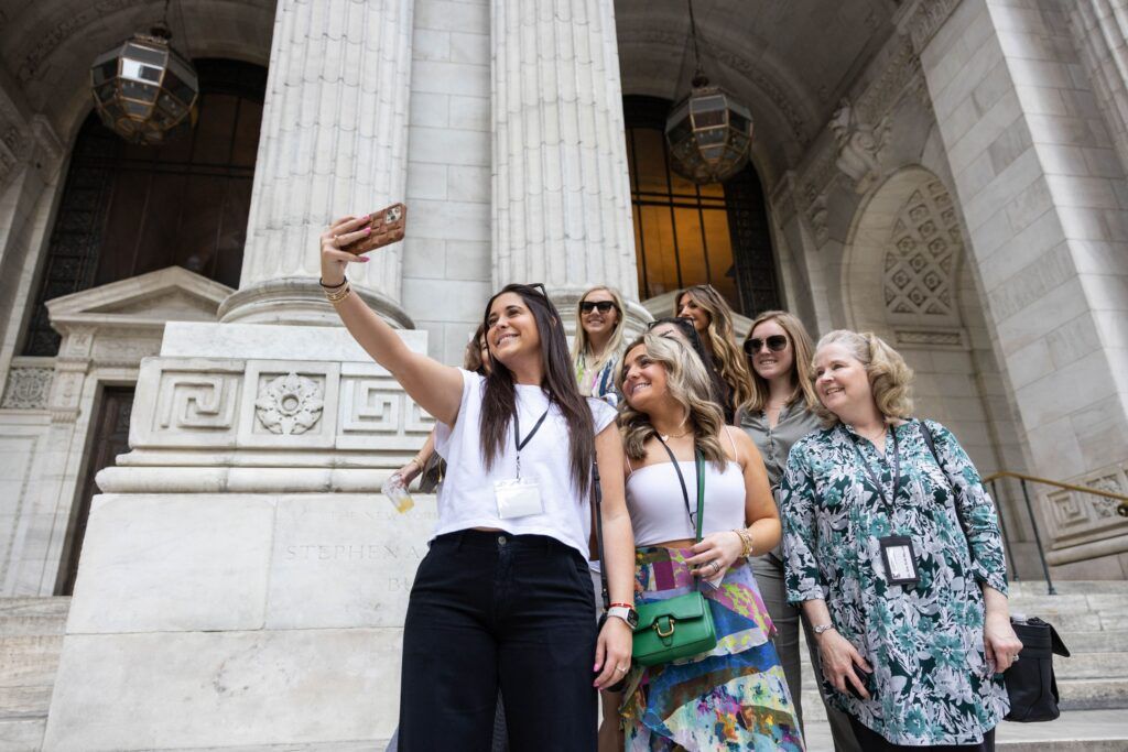 A young woman in a white top and black pants takes a photo of herself and six other women. The building behind them is light-colored and has archaic features.