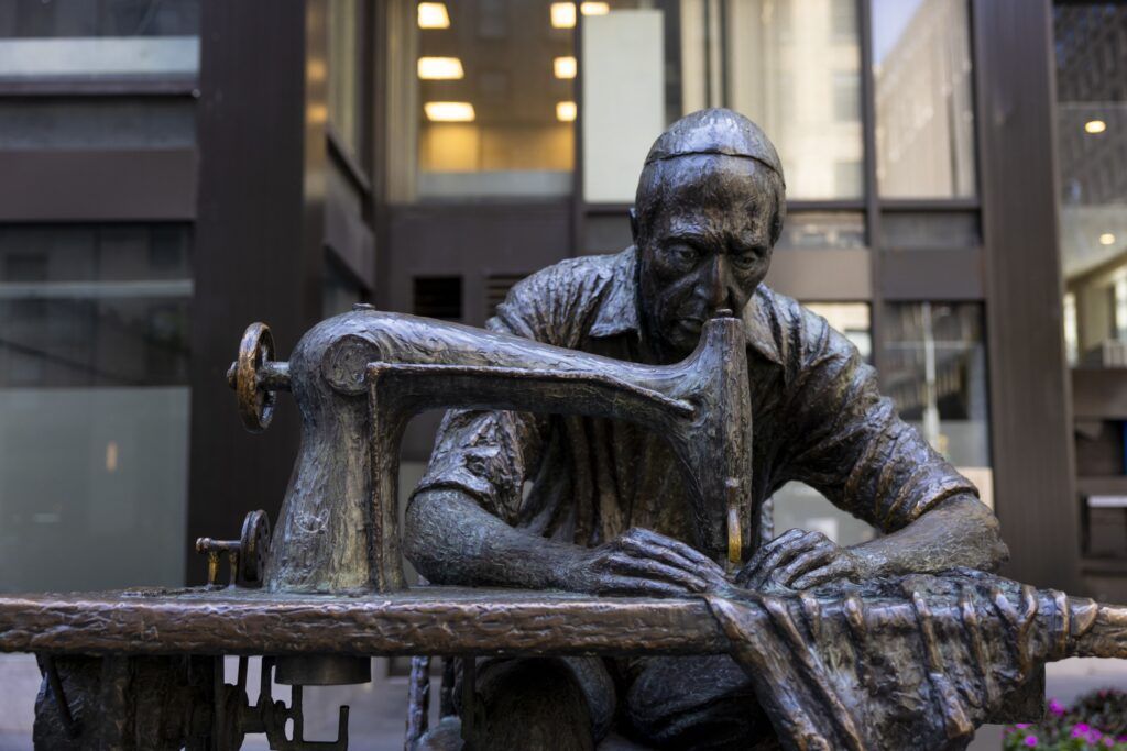 A dark metal statue of a man sewing cloth at a table with a sewing machine. There is a large glass and metal building in the background.