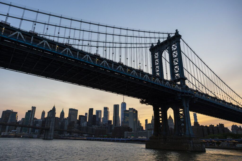 A closeup of a bridge over the water at sunset. There are many tall, dark buildings in the background with small lights coming through the windows.