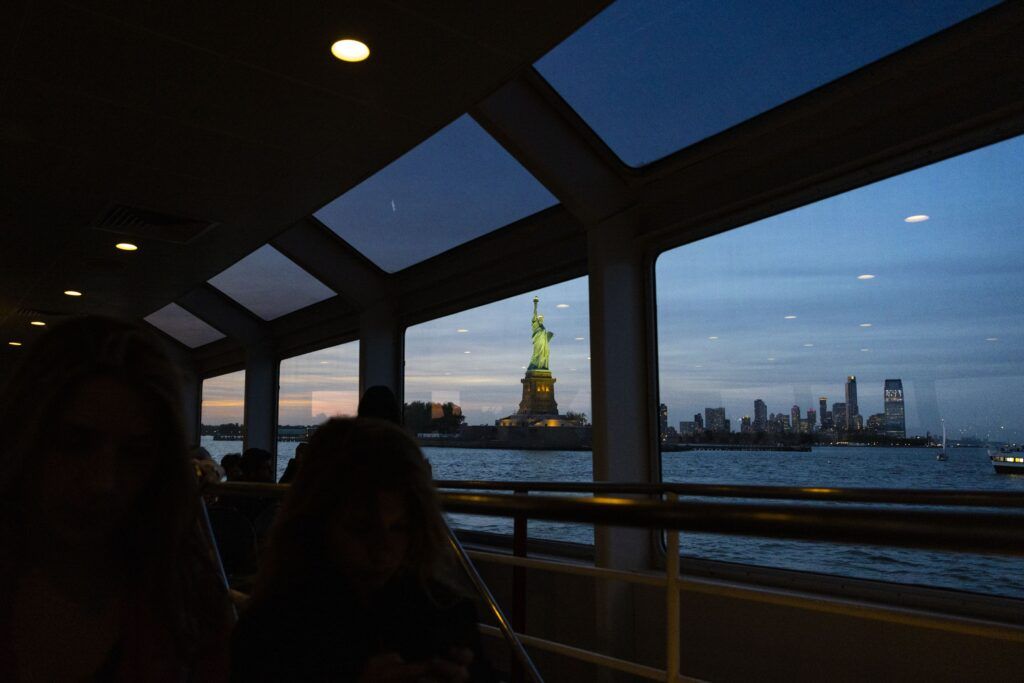 The view from inside a darkened boat. The glass windows look over the water and towards the Statue of Liberty. The lights from inside the boat reflect off the windows. There are other small boats and buildings in the distance.