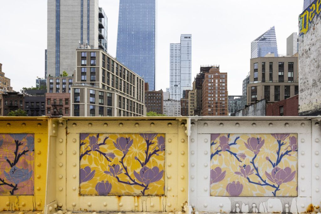 A paneled metal wall is decorated with purple stemmed flowers against varying yellow, oranges, and pink backgrounds. Behind the wall there are several tall buildings of various heights.