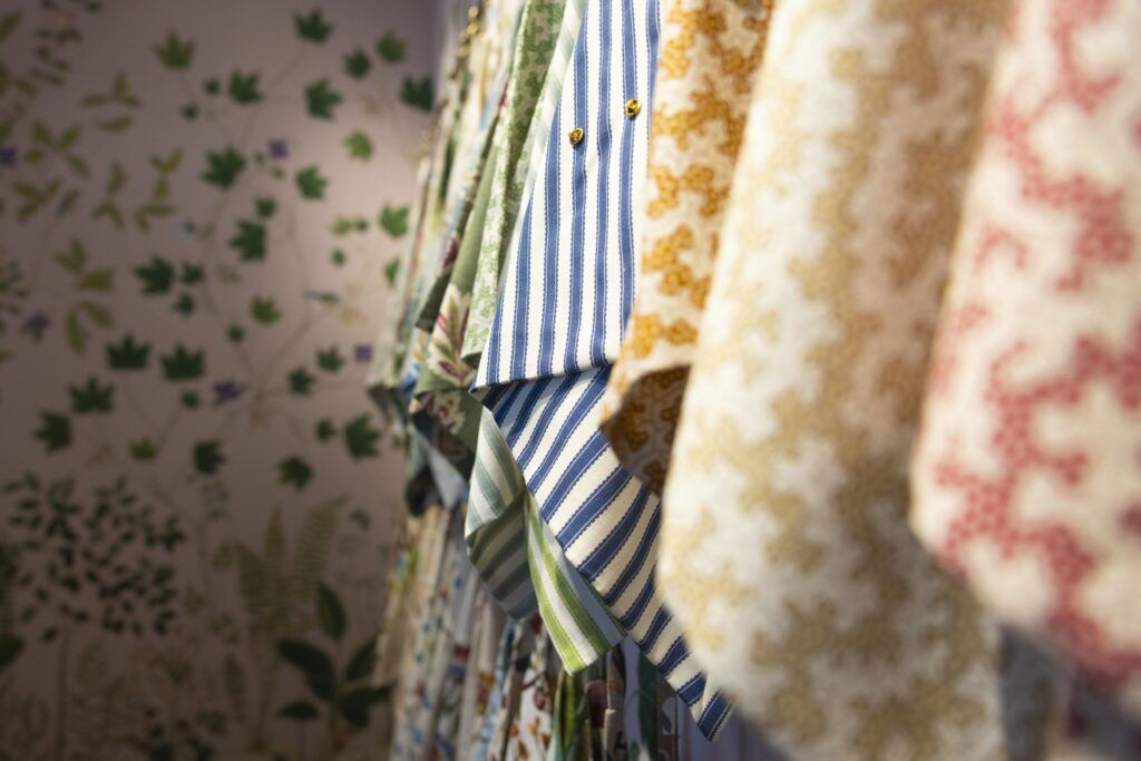 A closeup of several hanging fabrics of different colors and patterns. In the background is a wall patterned with green plants.