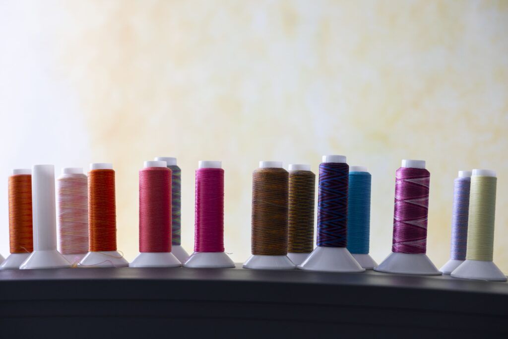 A closeup of 14 different spools of thread. The threads are different colors and patterns. The background is a yellow and white patterned wall.