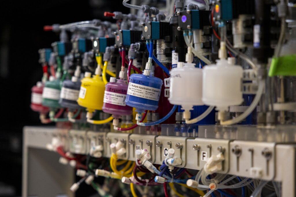 A closeup of the mechanics for a machine to dye fabric. There are several small plastic containers, each a different color, attached to tubes and labeled with singular letters.